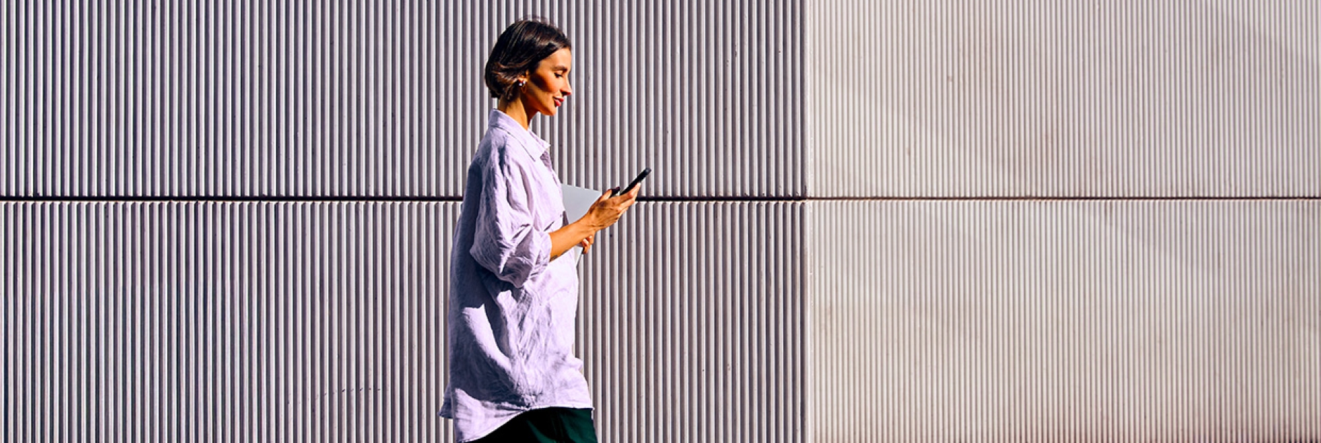 A person walks while looking at a mobile phone, with a corrugated metal wall in the background.