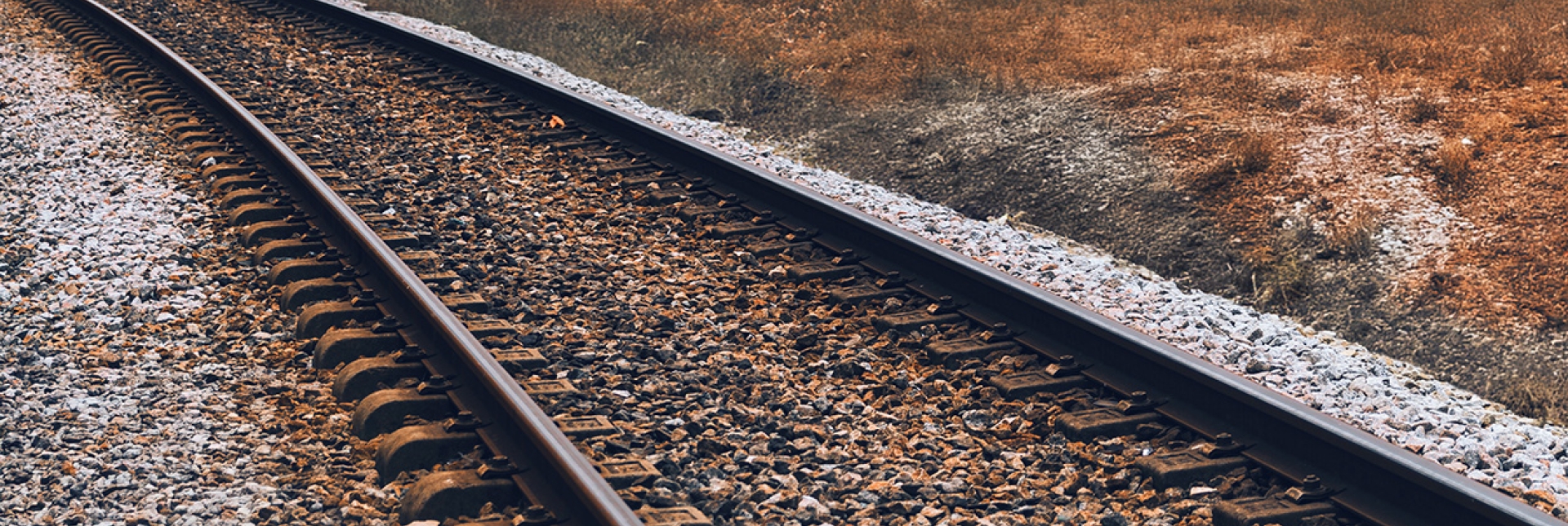 Curving railroad tracks run through a grassy field under an overcast sky.