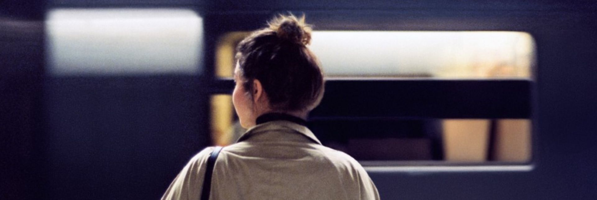 Woman with a beige jacket looking to her left as if she was waiting for a train, seen from the back