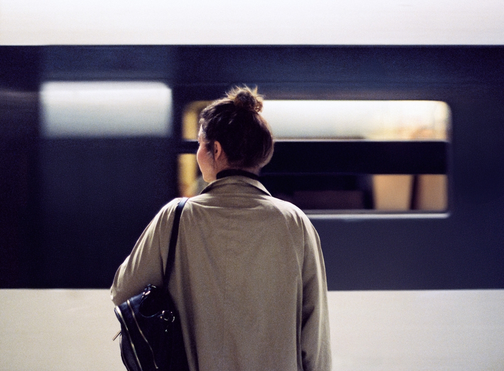 A person with their back to the camera stands in front of a train, wearing a light-colored coat and carrying a black shoulder bag. The person's face is pixelated