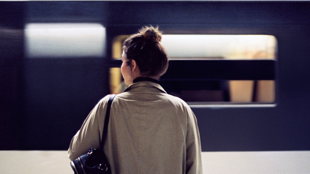 A person with their back to the camera stands in front of a train, wearing a light-colored coat and carrying a black shoulder bag. The person's face is pixelated