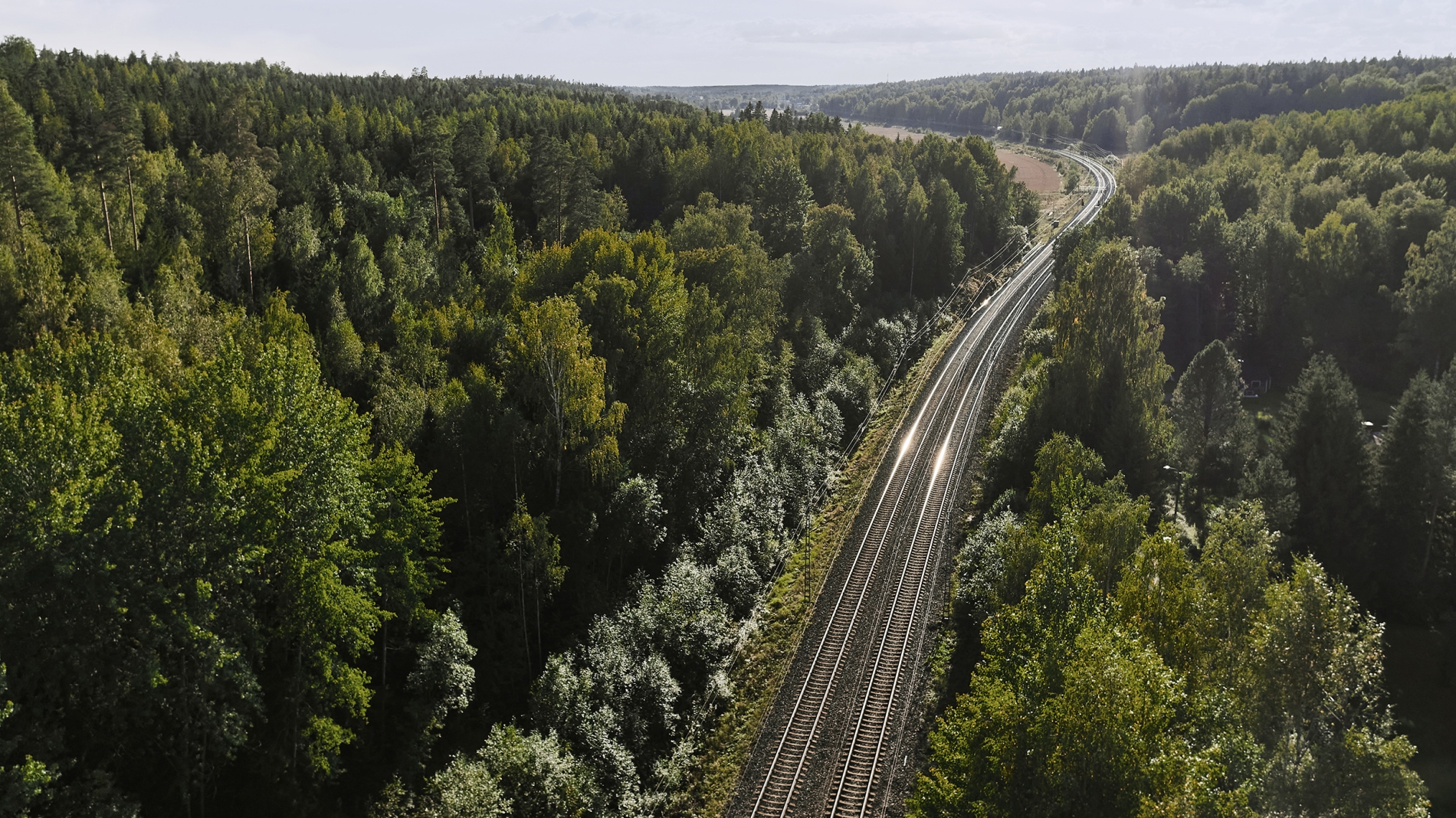 A railway track runs through a dense forest under a partly cloudy sky.