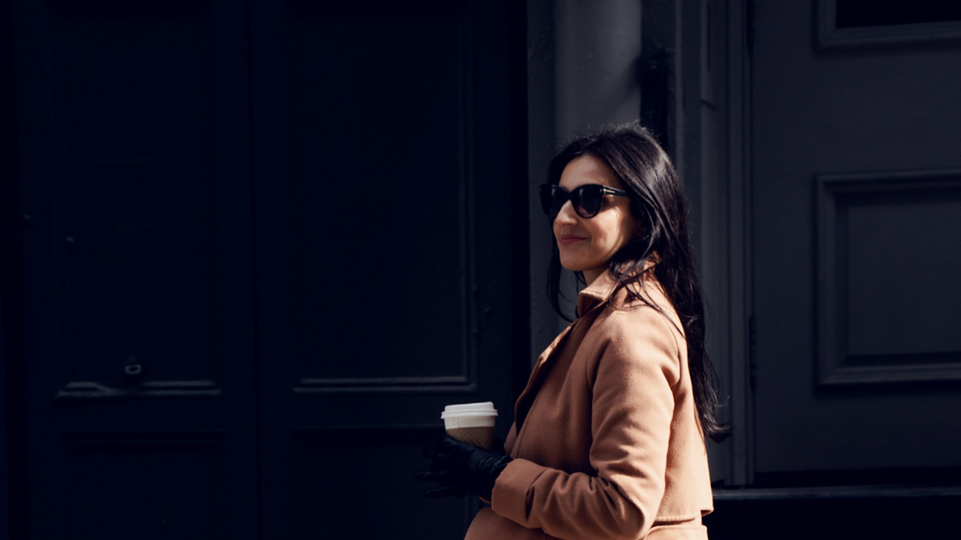 A person in a long brown coat walks down the street holding a cup.
