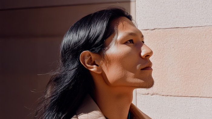 An Indigenous person in a beige jacket stands by a light-colored wall.