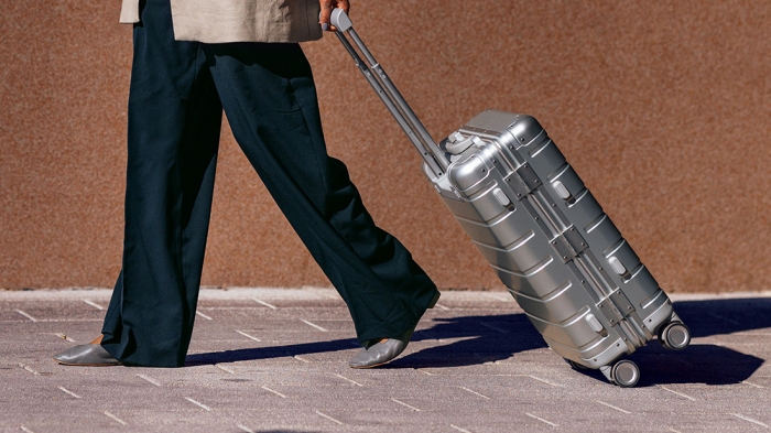 A person walks on a paved surface, pulling a silver suitcase past a brown wall.