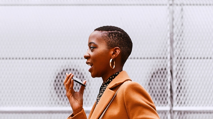 A person in a brown coat holds a smartphone near their face while carrying a black handbag. The background features a metal mesh fence with circular patterns