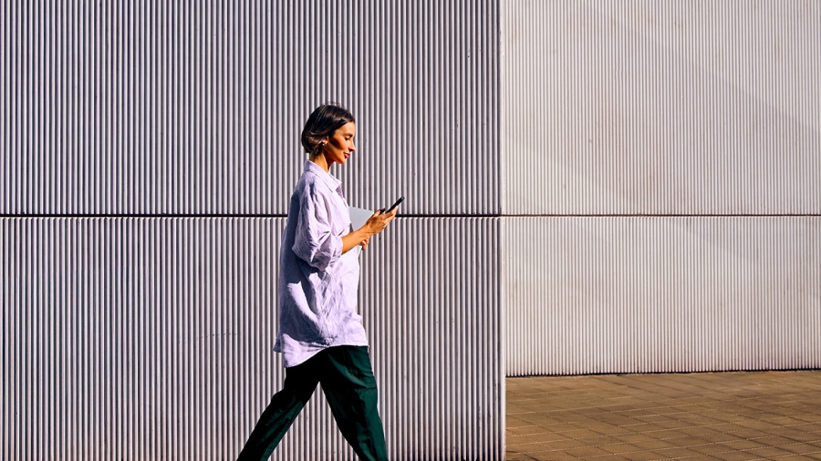 A person walks while looking at a mobile phone, with a corrugated metal wall in the background.