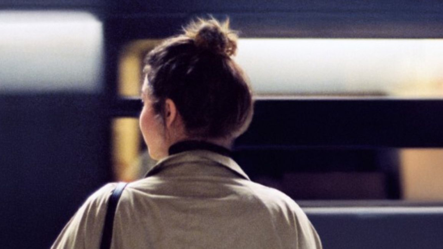 Woman with a beige jacket looking to her left as if she was waiting for a train, seen from the back