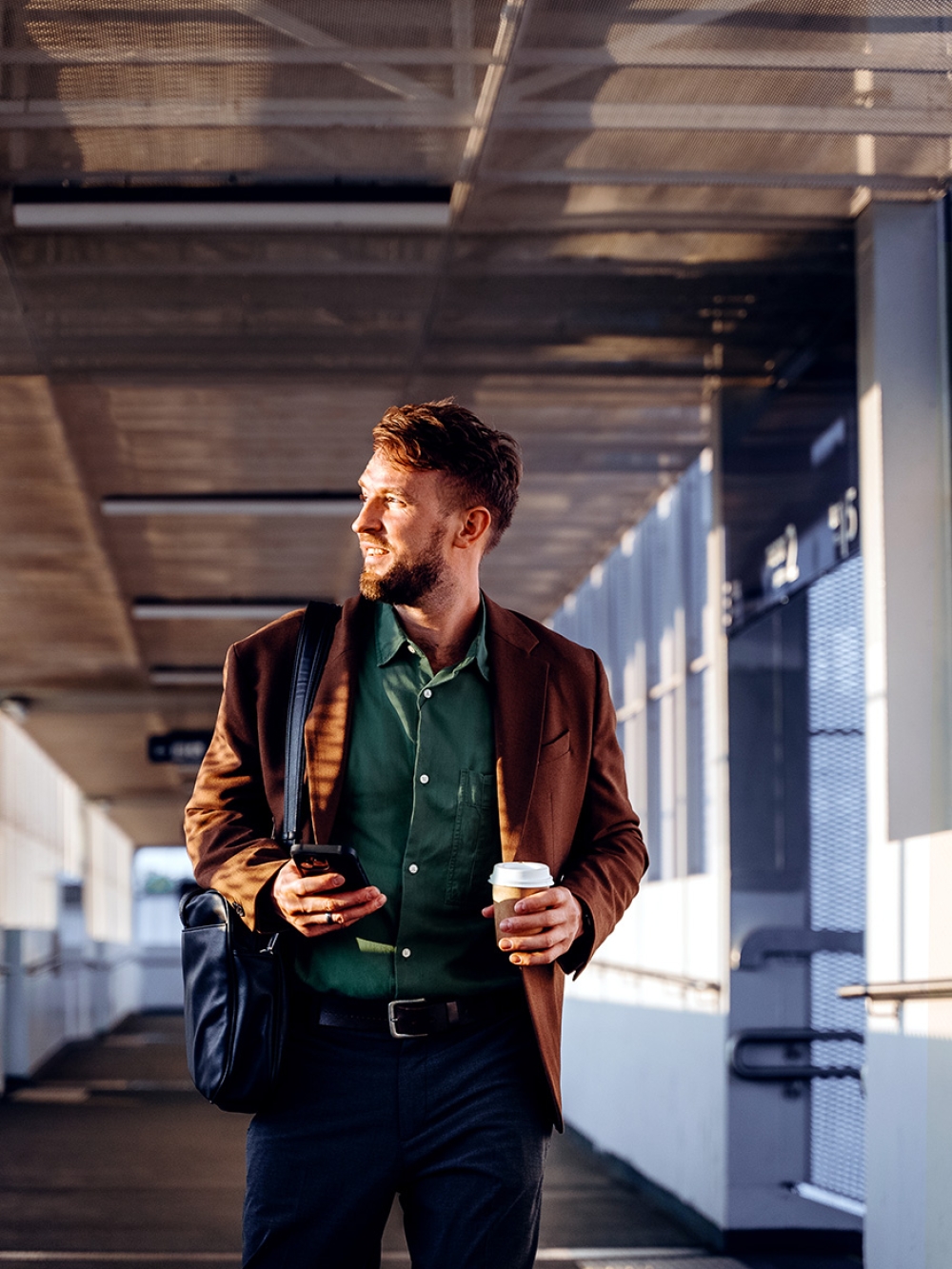 A person walks through a covered corridor, holding a smartphone and a coffee cup, with a black bag over their shoulder.