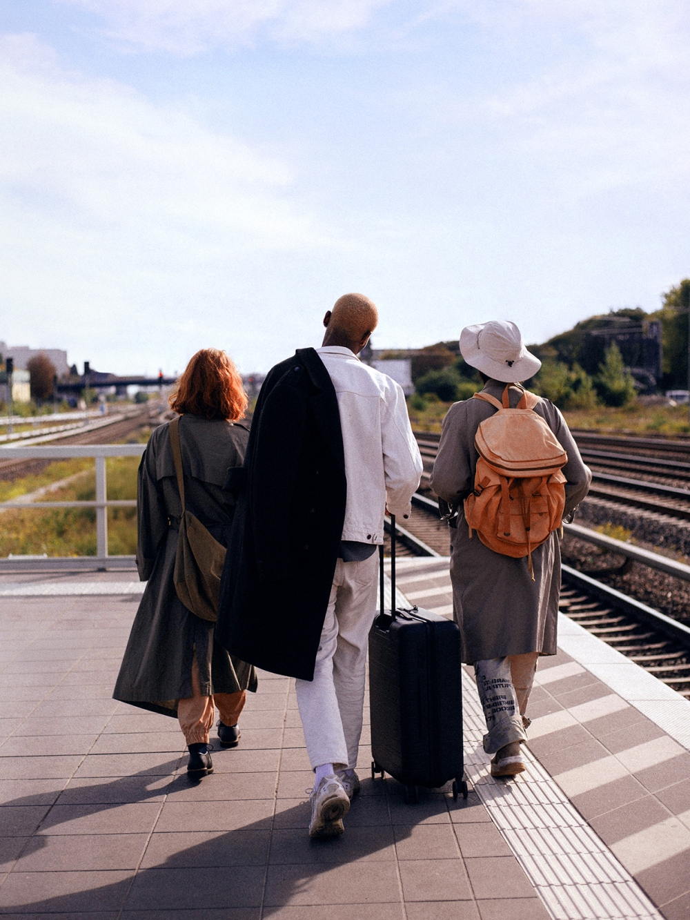 Three people walk along a train platform, two with coats and one pulling a suitcase.