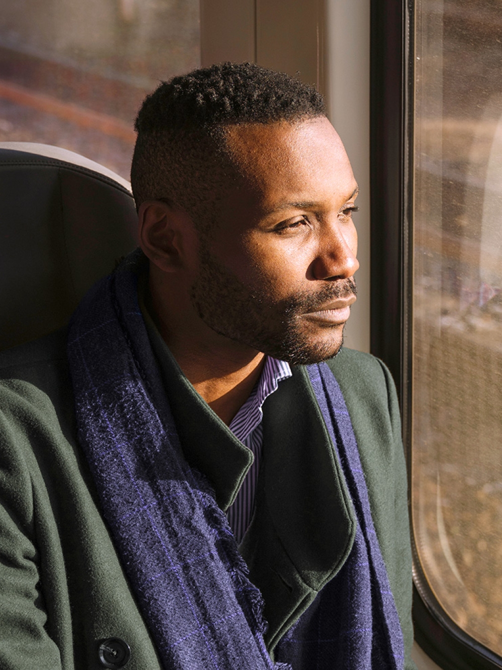 A person in a green coat and blue scarf sits by a train window.