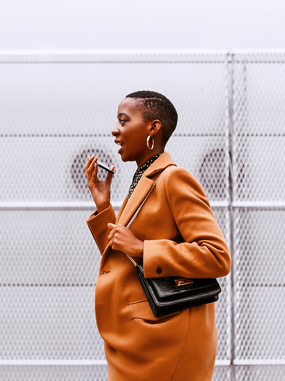 A person in a brown coat holds a smartphone near their face while carrying a black handbag. The background features a metal mesh fence with circular patterns