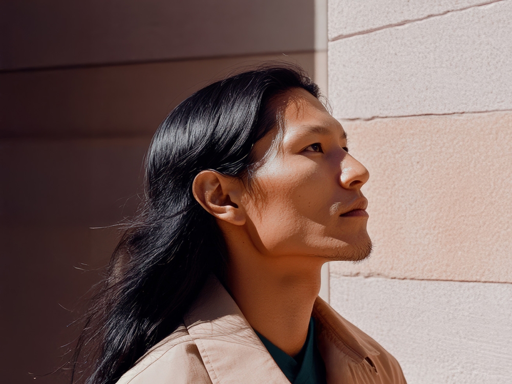 An Indigenous person in a beige jacket stands by a light-colored wall.