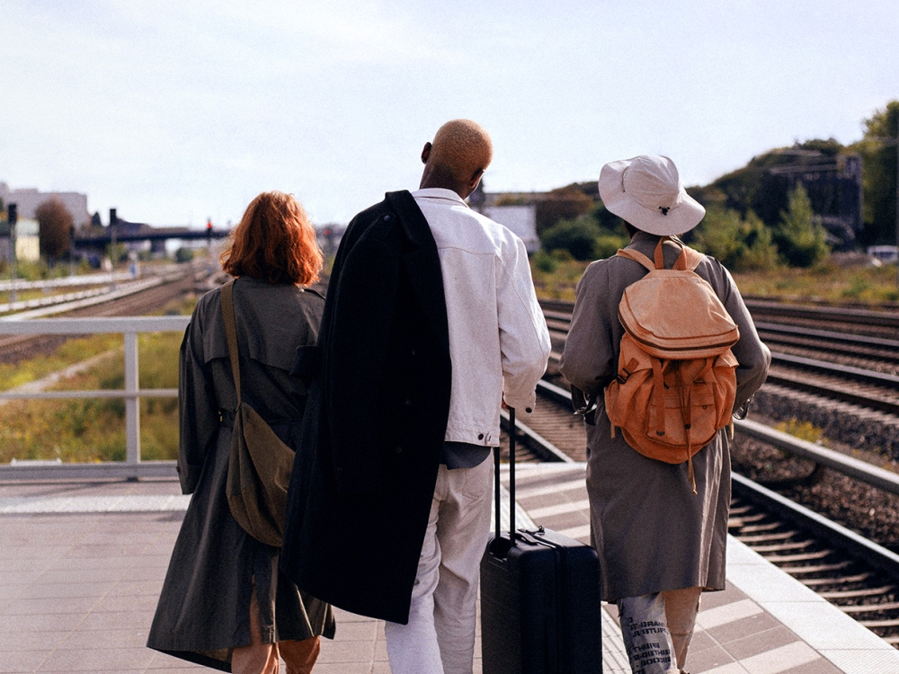 Three people walk along a train platform, two with coats and one pulling a suitcase.