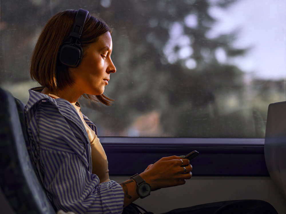 A person with headphones sits by a train window, holding a smartphone.